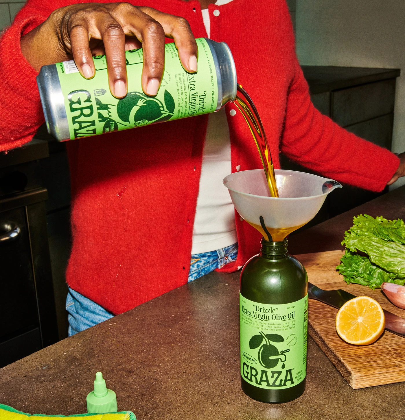 A hand pouring a Graza Drizzle refill can through a funnel into a squeeze bottle