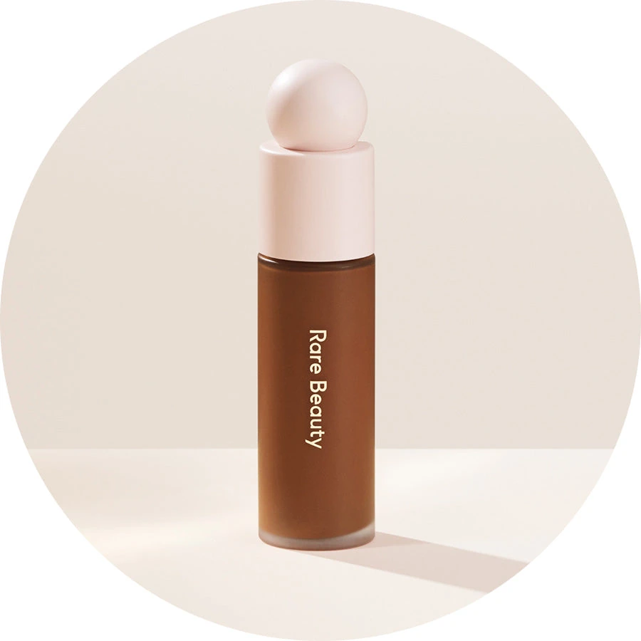 Rare Beauty Liquid Touch foundation bottle with a rounded cap in the brand’s beige studio setting