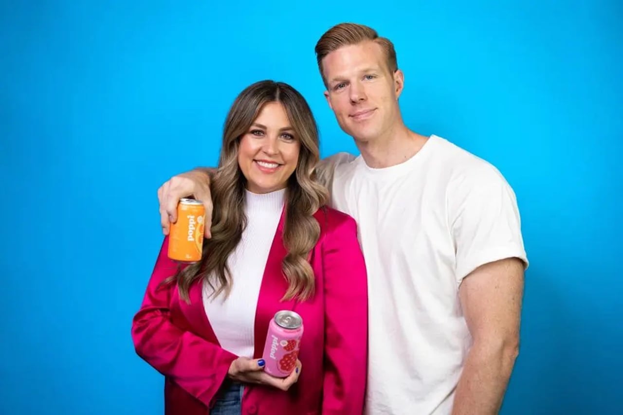 Poppi founders Stephen and Allison Ellsworth holding soda cans against a blue backdrop